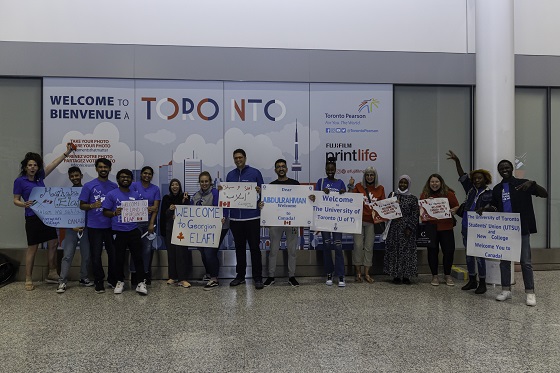Student refugee arrives at Toronto airport and is greeted by members of Team Georgian; group photo of people in airport holding up welcome signs and wearing Georgian blue
