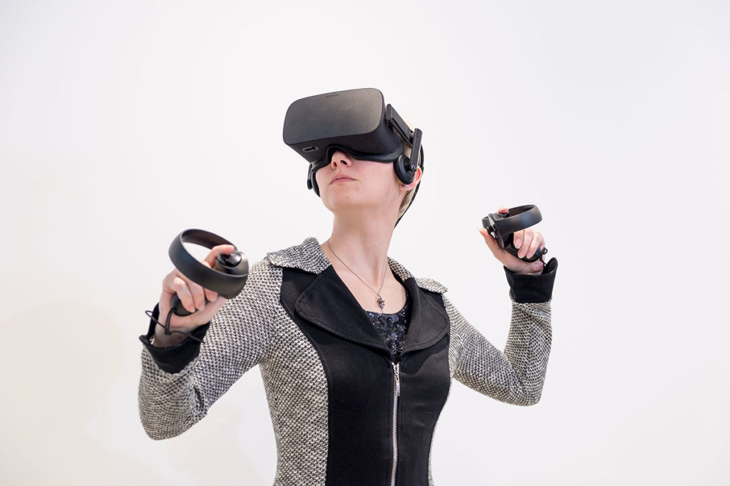 A young woman wearing a black and grey stripped shirt and black virtual reality goggles. She's holding controls in her hands.