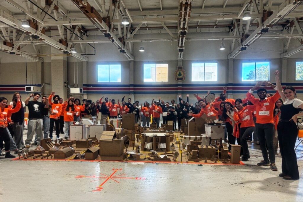 A large room with a group of people wearing matching orange and black shirts around displays made of cardboard sitting on the floor. 