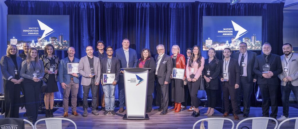 A group of people stand in a long row behind a podium and in front of screens reading "Mayor's Innovation Awards."