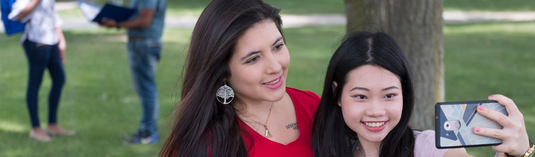 Two international students standing on the grass in front of a tree at Georgian College's Barrie Campus taking a selfie with a cellphone