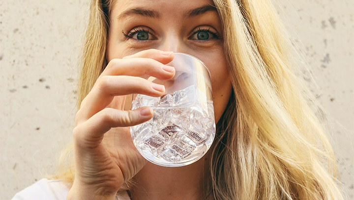 A person with blonde hair and blue eyes, drinking water out of a glass with ice cubes