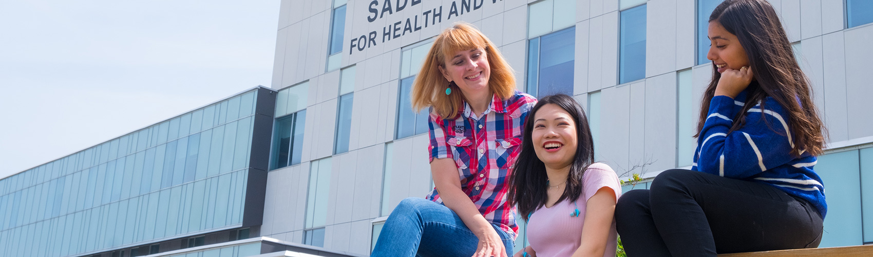 Three female international students sitting on a picnic bench in front of the Sadlon Centre for Health and Wellness (M building) at the Barrie Campus