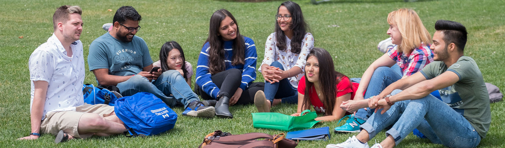 A group of eight international students sitting on green grass with backpacks and binders around them