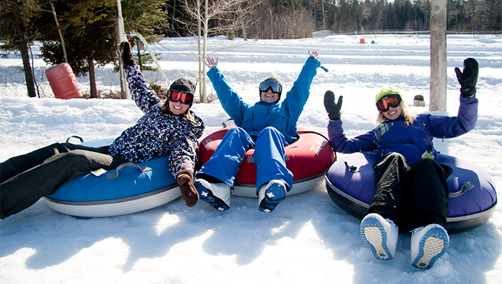 A group of people in winter boots, snow suits, goggles and gloves, sitting in snow tubes with their arms up in the air at Snow Valley