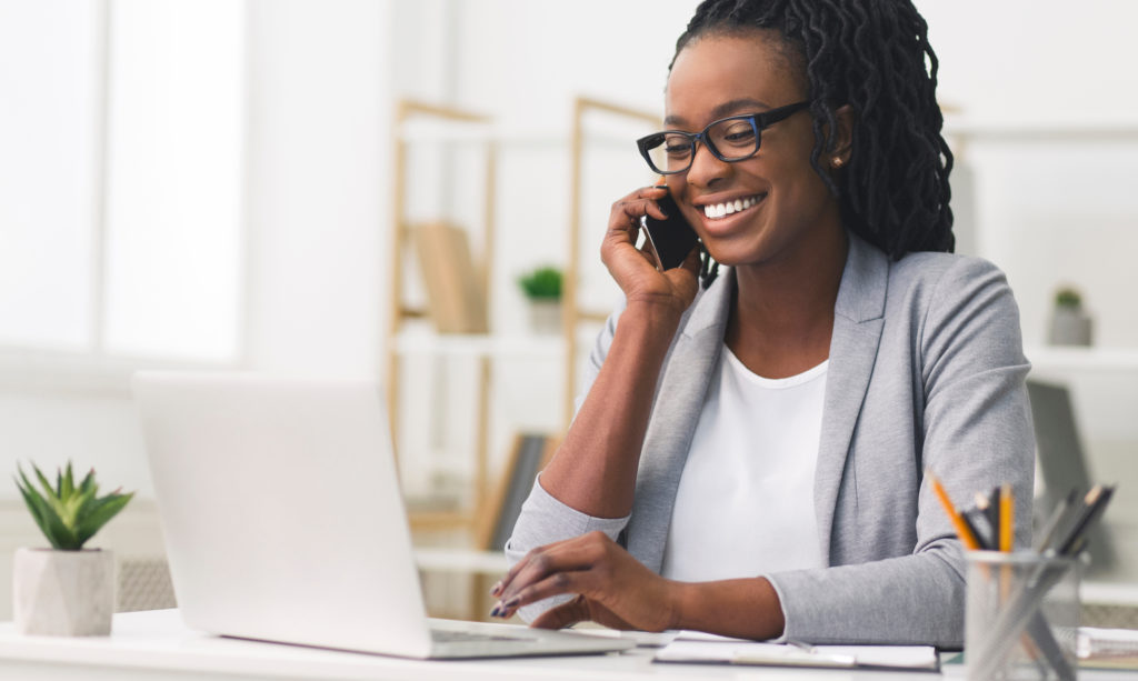 A person with dark hair smiling while holding a cellphone to their ear in an office setting