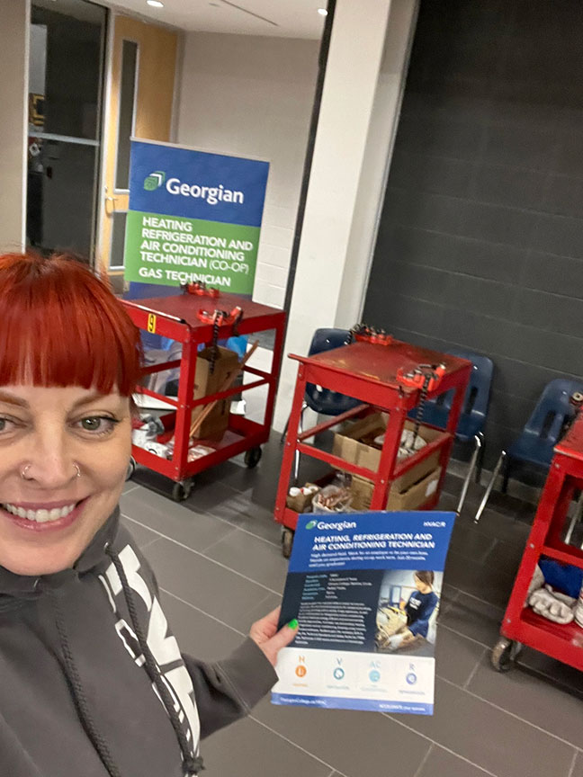 A smiling female with red hair (Brandi Ferrenc) holding up a flyer and taking a selfie. She's standing in front of red carts and a banner.