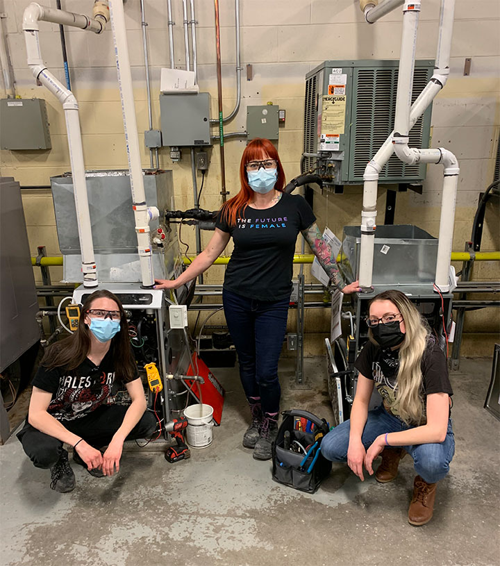 three females wearing face masks (one is Brandi Ferenc) in front of hvac equipment. The middle female is standing and the other two are squatting.