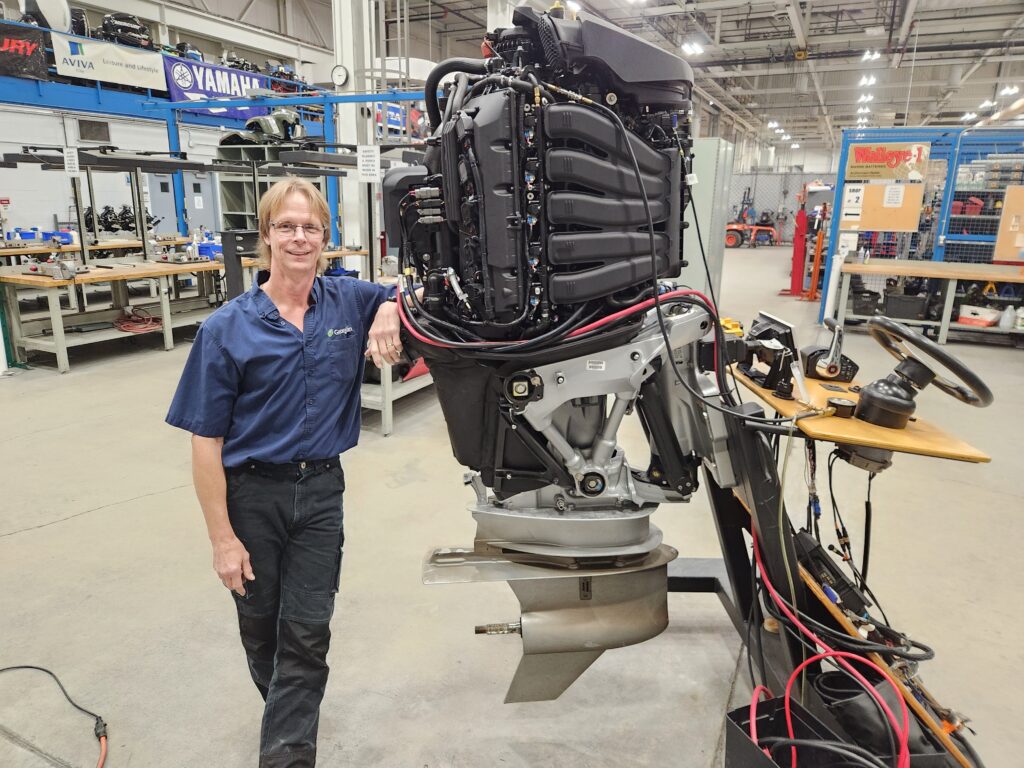 A person stands in a workshop and leans an arm against an oversized boat motor. 