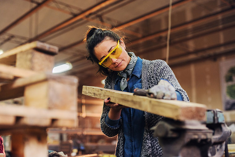 A female with long dark hair holding a piece of wood in a woodshop. She's wearing safety glasses.