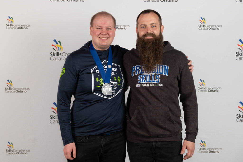 Two people, one of whom has a silver medal around their neck, stand in front of a backdrop reading "Skills Ontario."