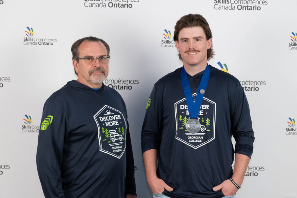Two people, one of whom wears a silver medal around their neck, stand in front of a backdrop reading "Skills Ontario."