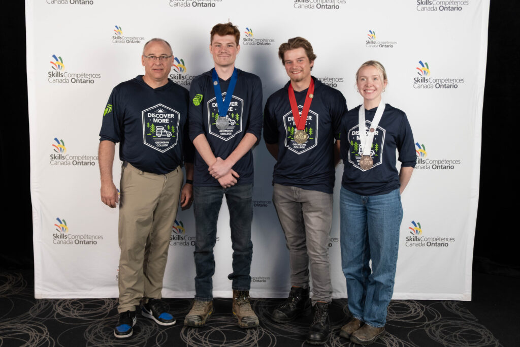 Four people, three of whom are wearing medals around their necks, stand in front of a backdrop reading "Skills Ontario."