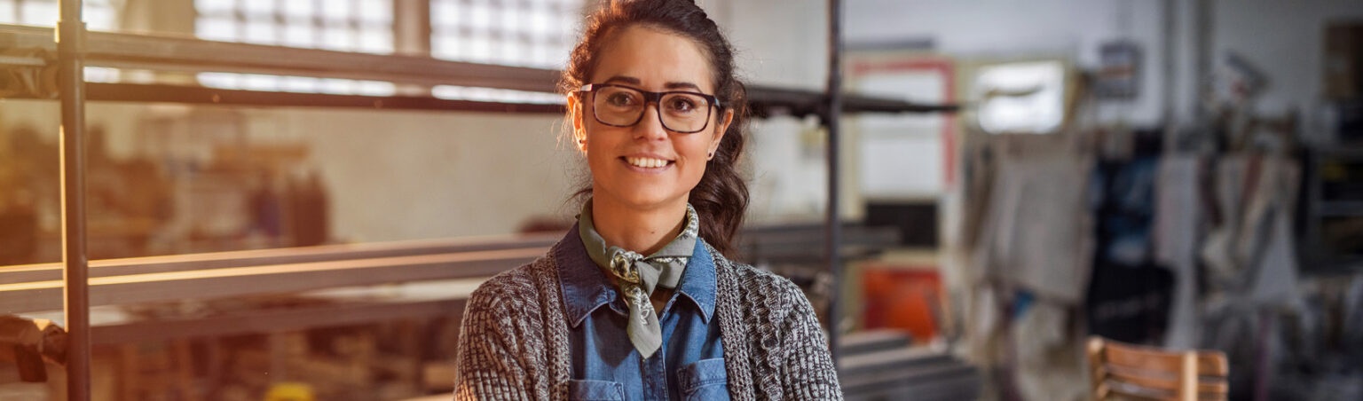 A person with brown hair pulled back in a ponytail, black large-framed glasses, wearing a denim collared button-up shirt with a grey knit cardigan and a sage green neck scarf, standing in a workshop smiling with their arms crossed