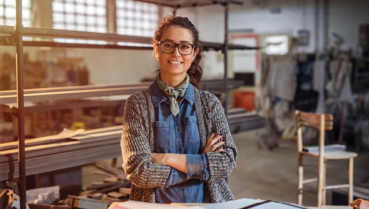A person with brown hair pulled back in a ponytail, black large-framed glasses, wearing a denim collared button-up shirt with a grey knit cardigan and a sage green neck scarf, standing in a workshop smiling with their arms crossed