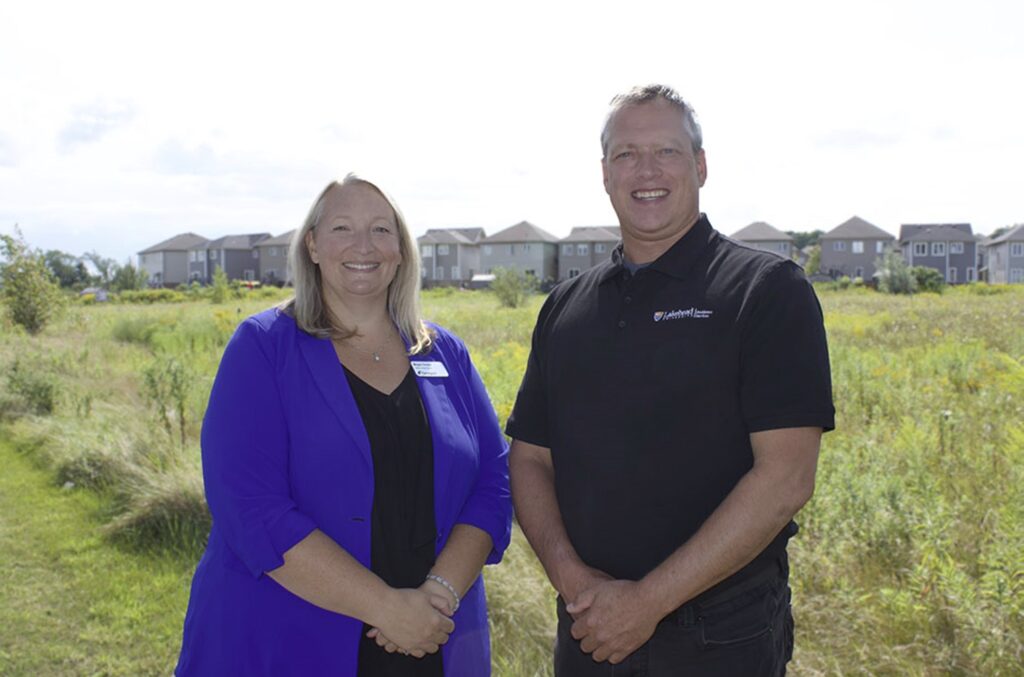 Two people stand in front of a row of houses in the background. 