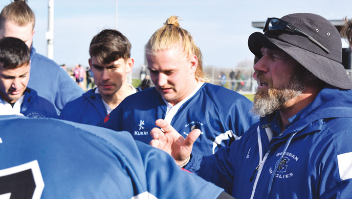 Georgian Grizzlies coach speaking to a team of rugby players in uniforms mid-game on a sports field
