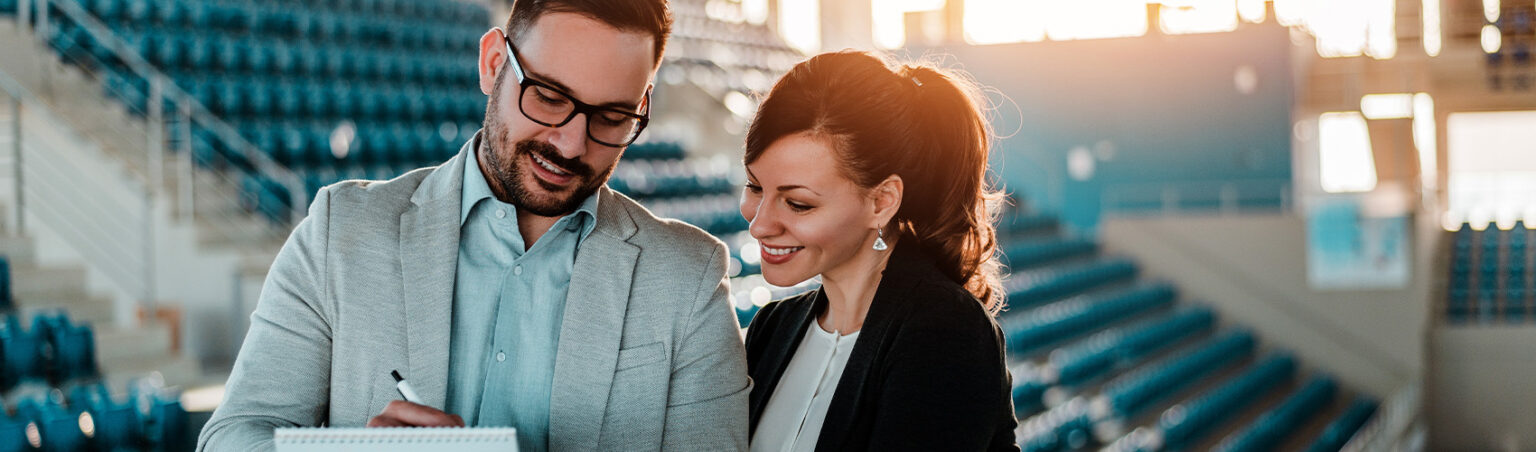 Two professionals standing in a sports facility with rows of blue seats.