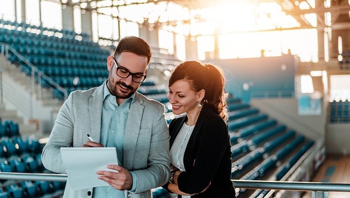 Two professionals standing in a sports facility with rows of blue seats.