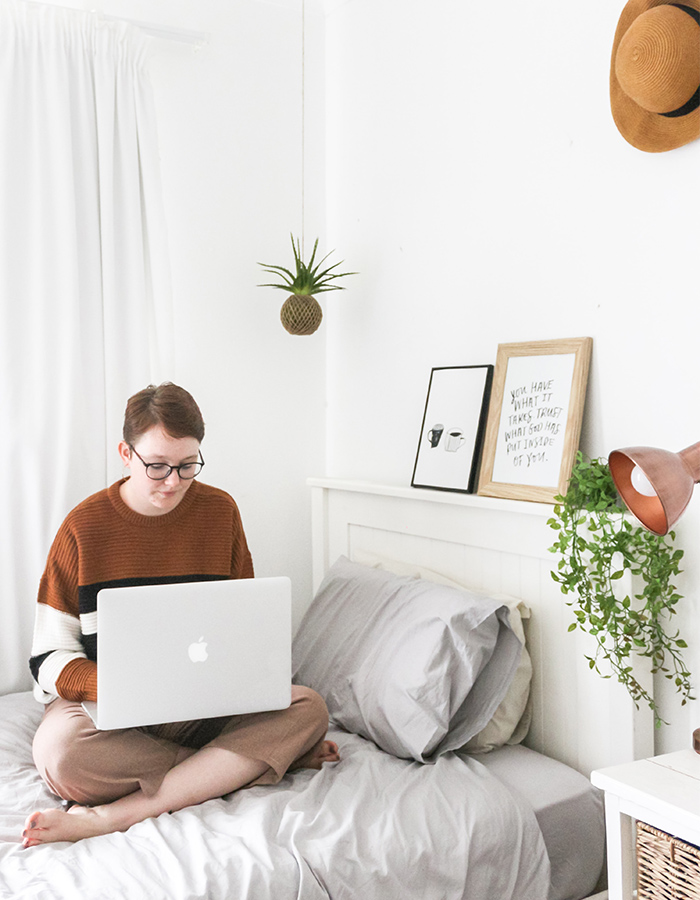 person with short hair and glasses sitting cross-legged on a bed using a Mac laptop