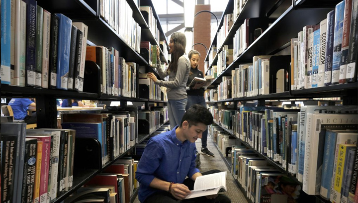 Three students standing between the book shelves at the library reading