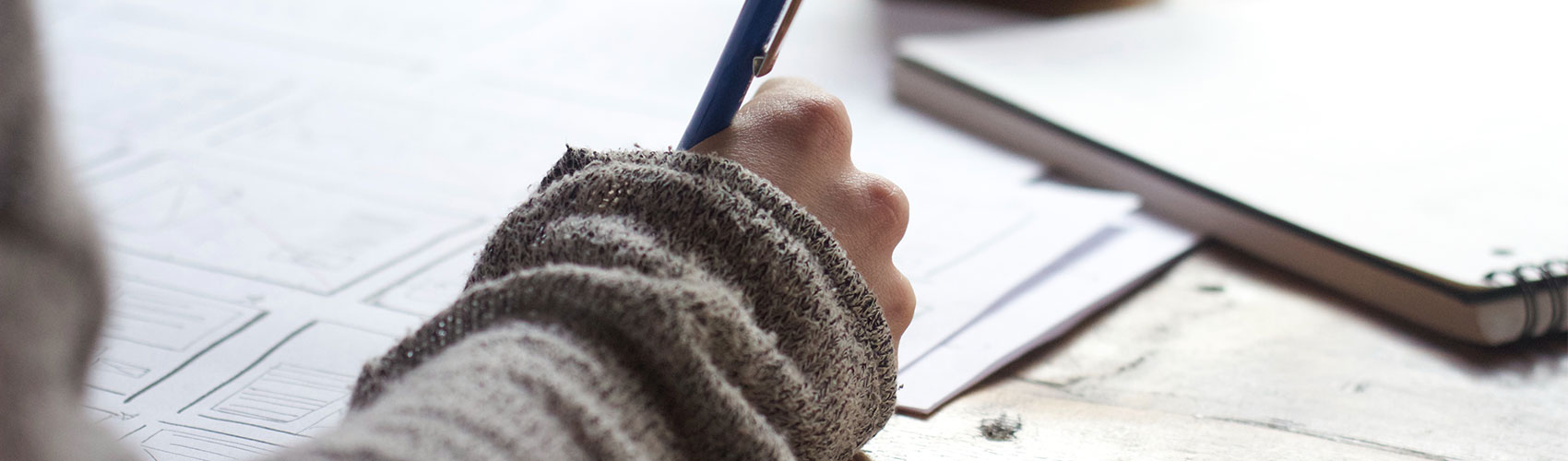 person wearing beige, knit sweater holding a blue pen and writing on paper on a wood desk