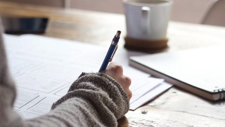 student holding a pen writing on paper with a coffee mug, notebook and phone on the table