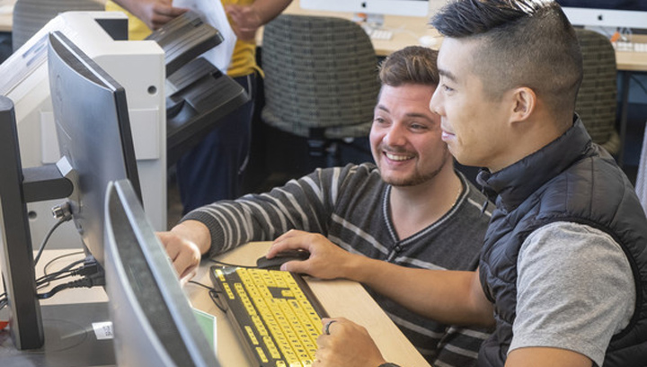 a male student crouching down next to a student working on a computer in the library