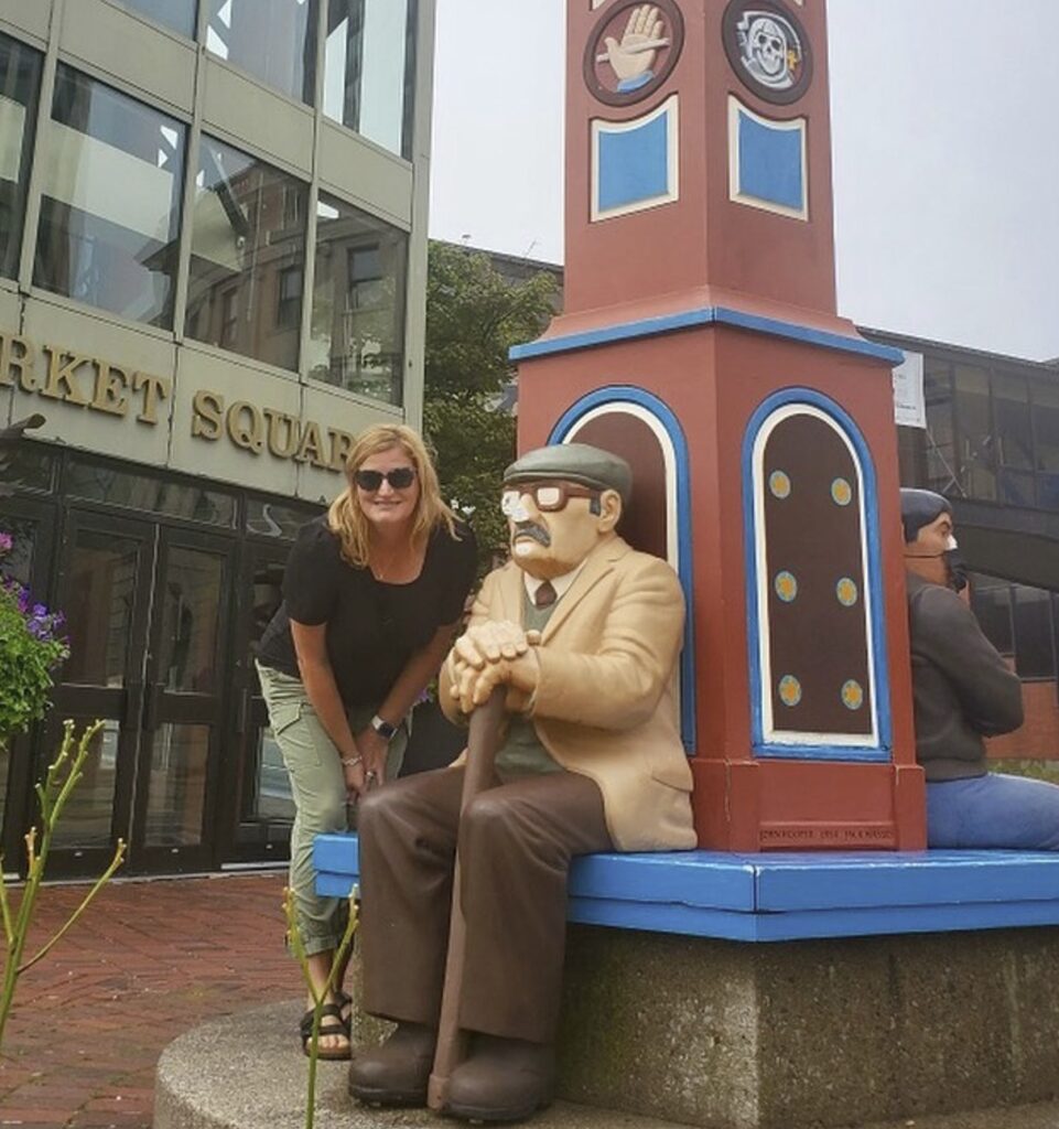 A person stands next to a statue of an old man sitting down and holding a cane.