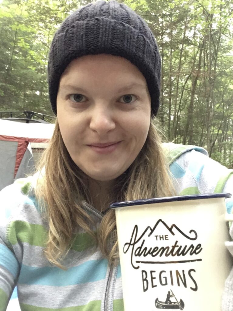 A person takes a selfie while sitting outside and holding a mug reading "The Adventure Begins."