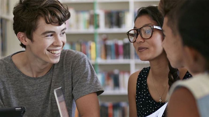 Student with short brown hair on the left side of the photo is smiling and engaging with another student and instructor in a library. 