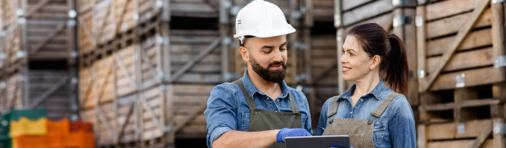 Two workers wearing denim shirts and overalls stand in front of stacked wooden crates.