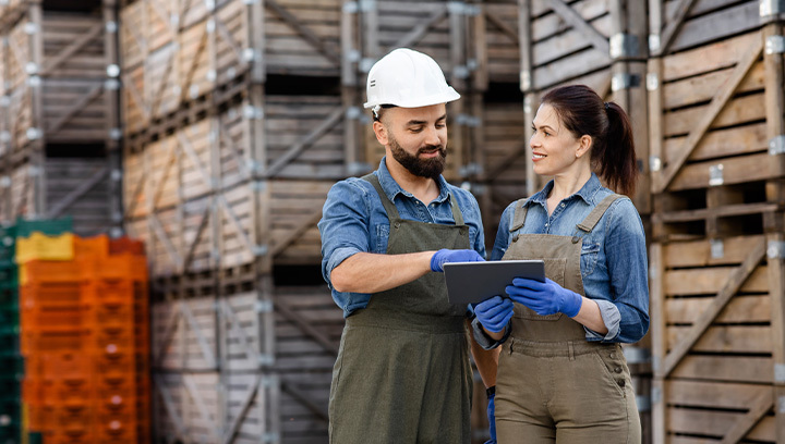 Two workers wearing denim shirts and overalls stand in front of stacked wooden crates.