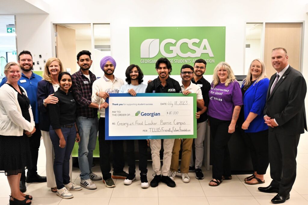 A group of people stand together in front of a sign reading "GCSA" and hold up an oversized cheque for $10,000.