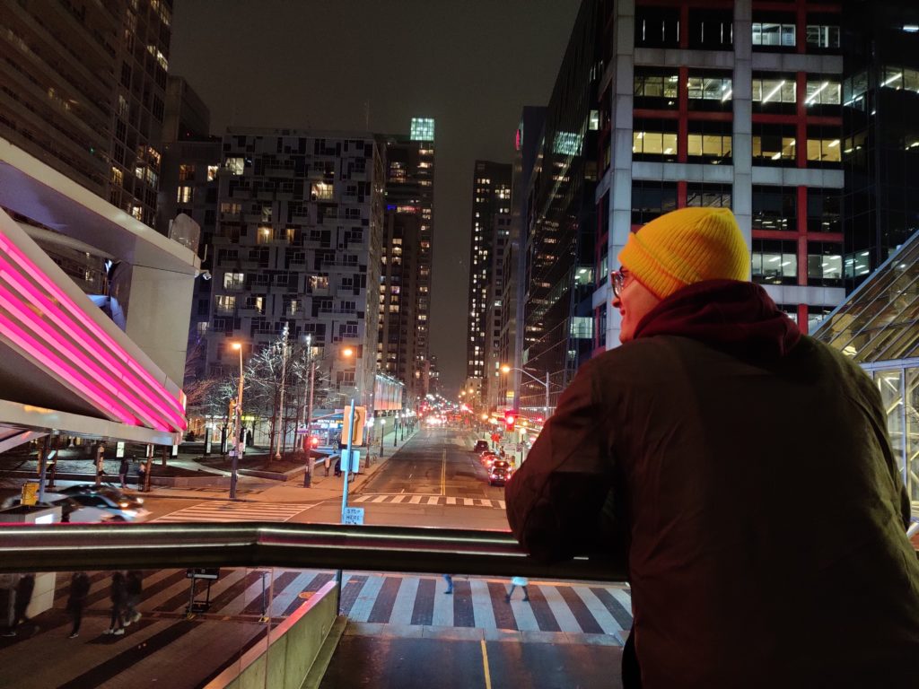 A person wearing a black jacket, yellow toque, and glasses smiles while leaning over a railing that overlooks a city at night.