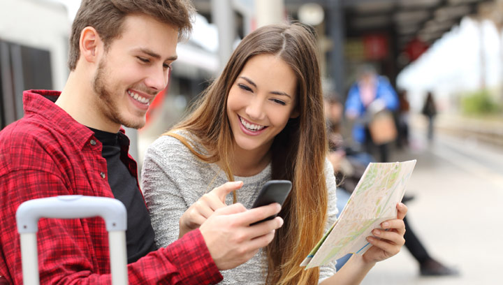 Two Tourism students smiling and sitting at a train station next to luggage while holding a cellphone and paper map