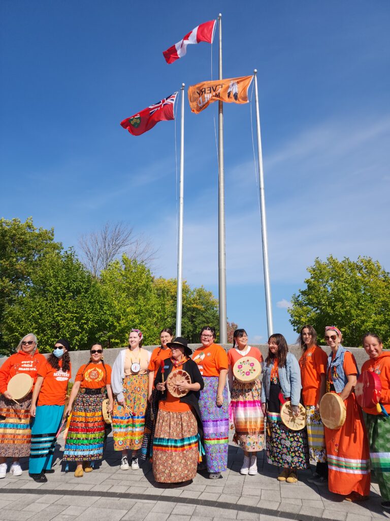 A group of people wearing orange shirts and ribbon skirts and holding drums stand together in front of three flagpoles. 