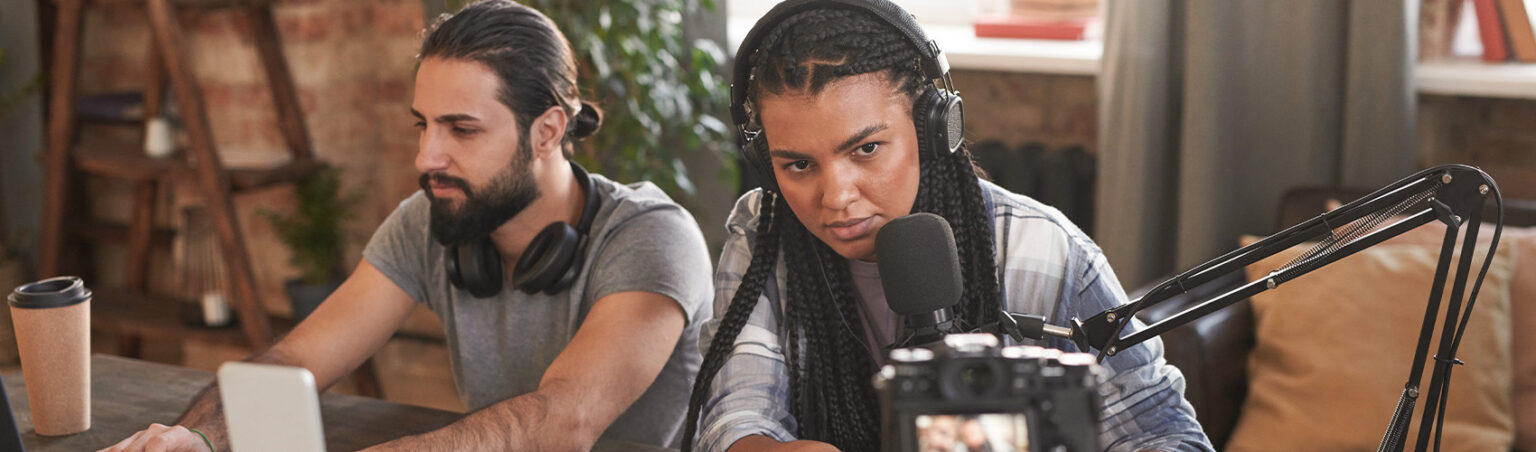 Two people working in a creative studio, one using a laptop and the other speaking into a microphone with headphones on, recording content.