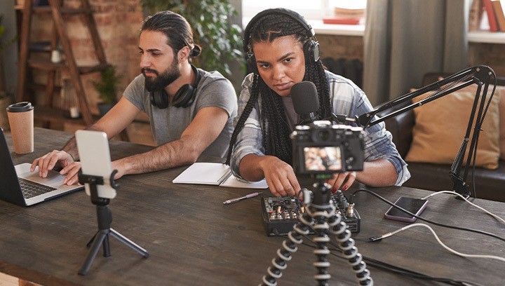Two people working in a creative studio, one using a laptop and the other speaking into a microphone with headphones on, recording content.